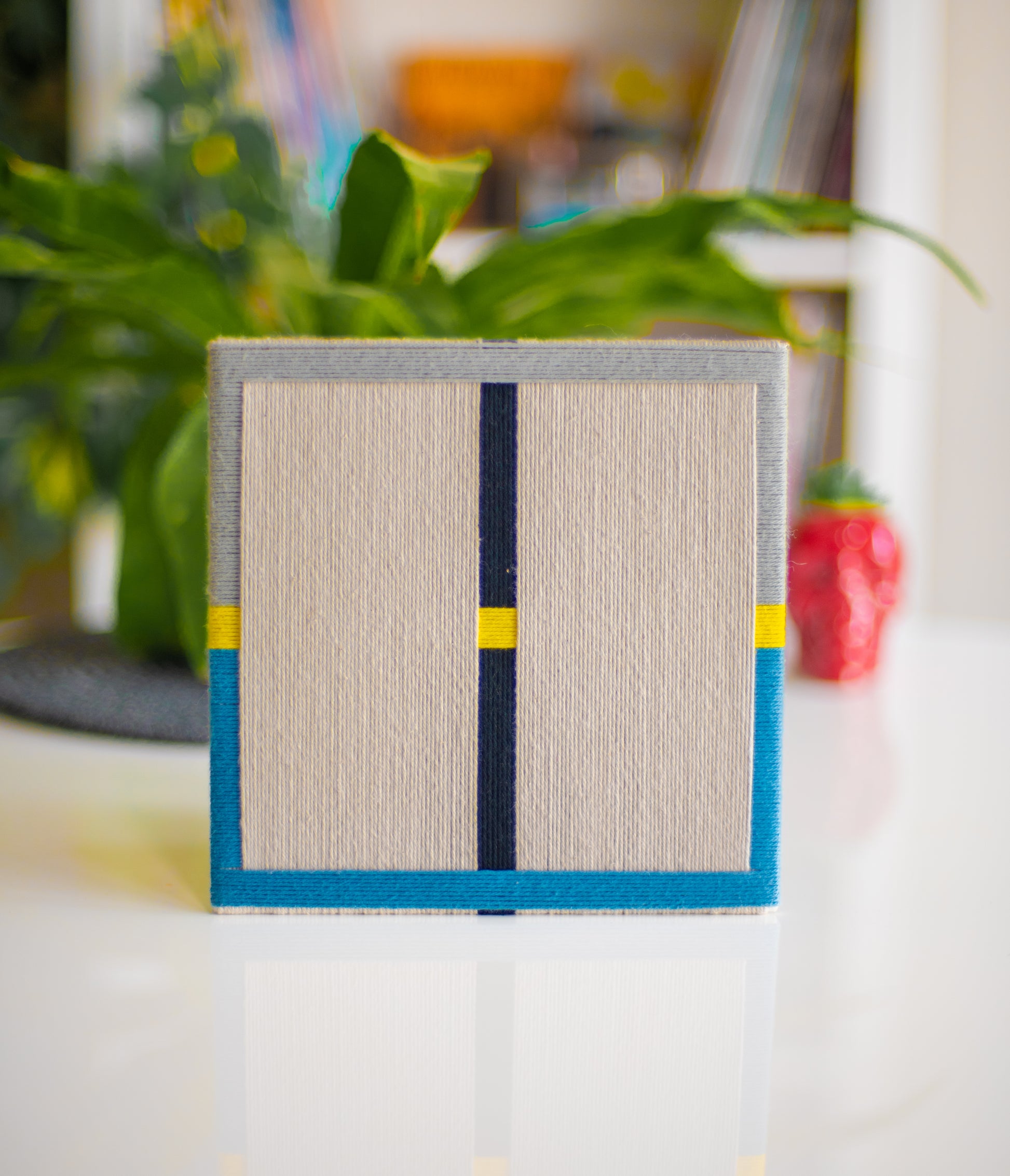 A rectangular textile wall art made from 100% cotton, featuring a neutral color with geometric patterns in black, blue, and yellow, displayed on a white surface with a plant in the background.