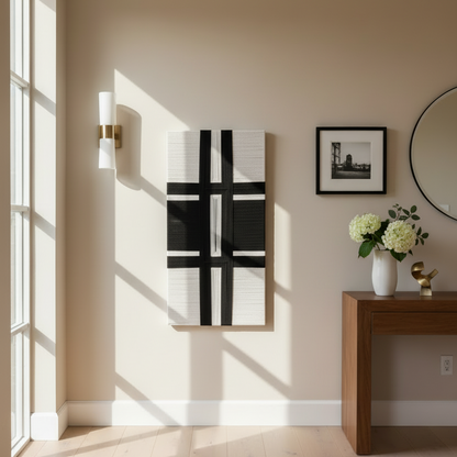 Modern interior with a black and cream abstract textile art, a vase with flowers, and a mirror on a beige wall.
