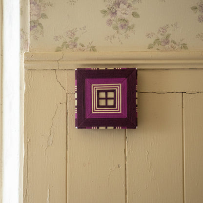 A square textile art in purple and chartreuse. Mounted on a cream paneled wall with floral wallpaper above.